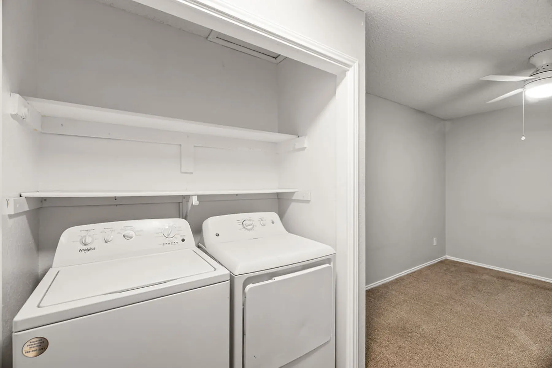 Laundry Room Setup A laundry room featuring a washing machine and dryer side by side, with white shelves above them and an adjoining room visible in the background.
