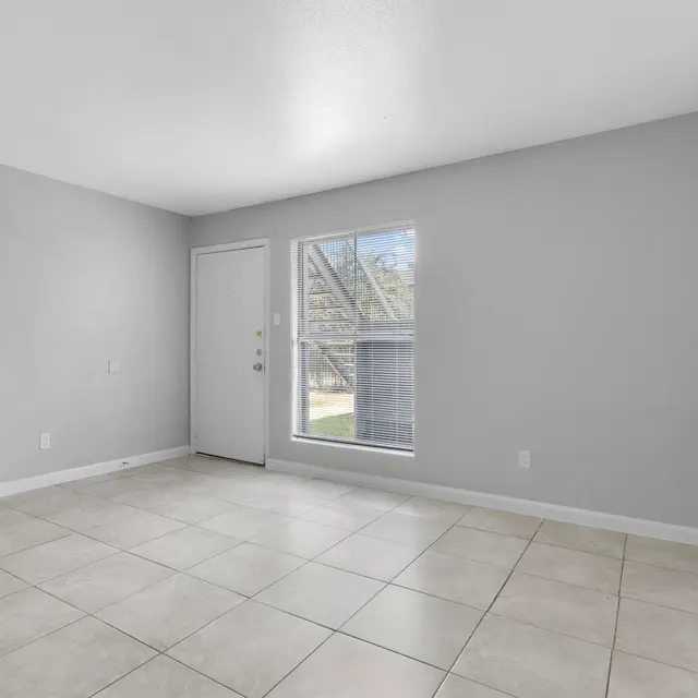 A spacious empty room with gray walls and tile flooring. A door on the left and a window on the right allowing natural light in.