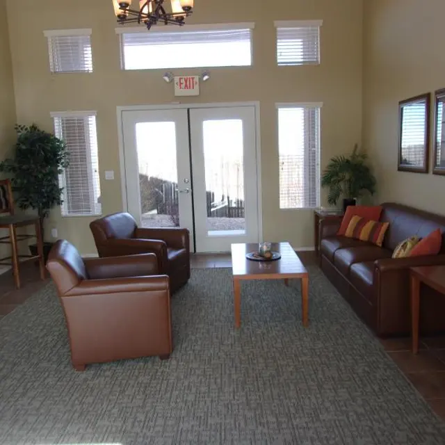 Cozy Living Room Design A cozy and modern living room with brown leather furniture, including a sofa and two armchairs, arranged around a small coffee table. Large windows let in natural light, and there are decorative plants and framed pictures on the walls. The flooring is tiled with a textured rug underneath the coffee table.
