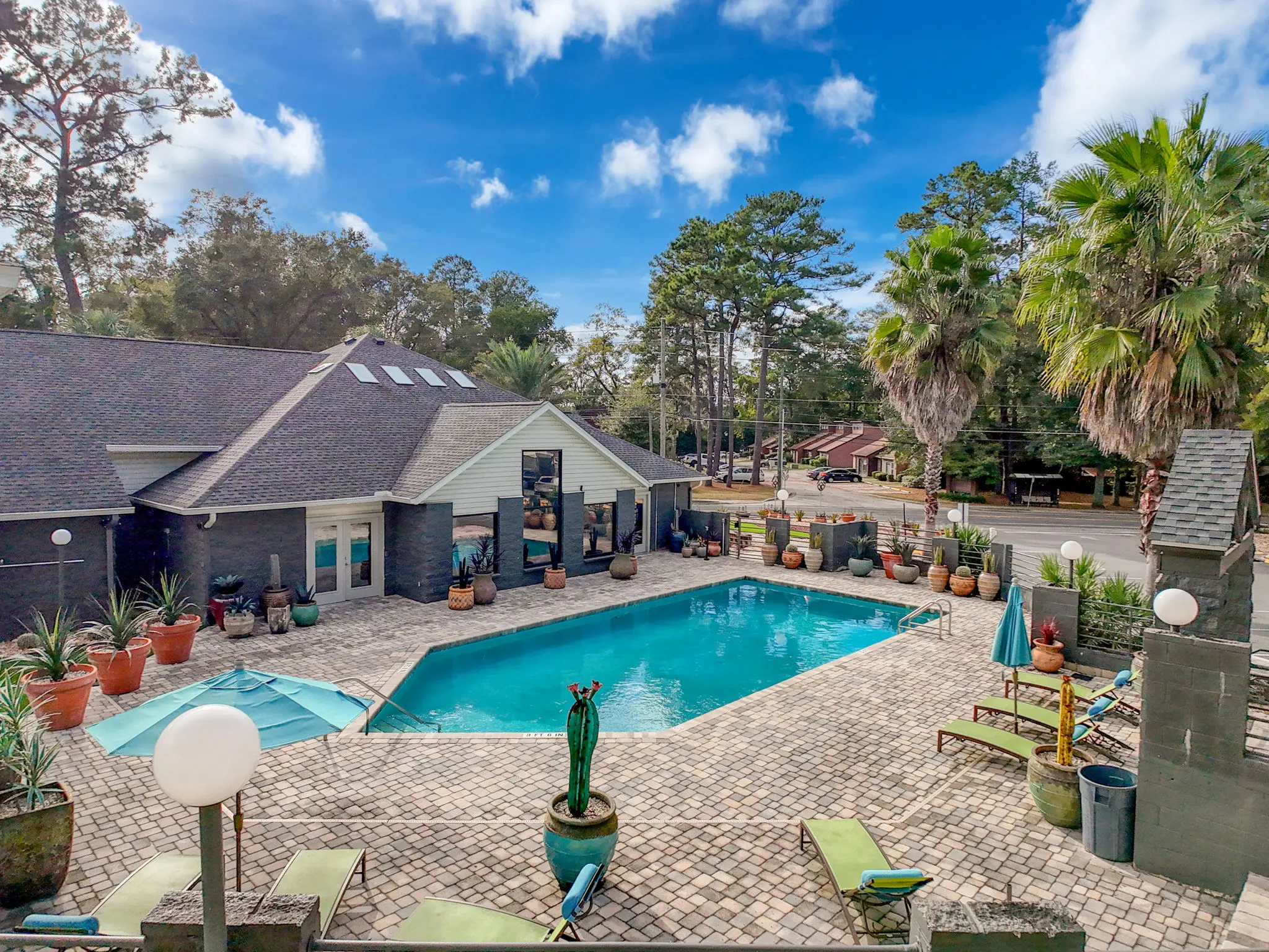 A spacious outdoor pool area surrounded by lounge chairs and potted plants, with a building in the background and a clear blue sky above.