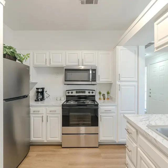A modern kitchen with white cabinets, stainless steel appliances including a refrigerator, stove, and microwave, and a light wood floor. A sink is visible on the right, and a coffee maker is placed on the counter.