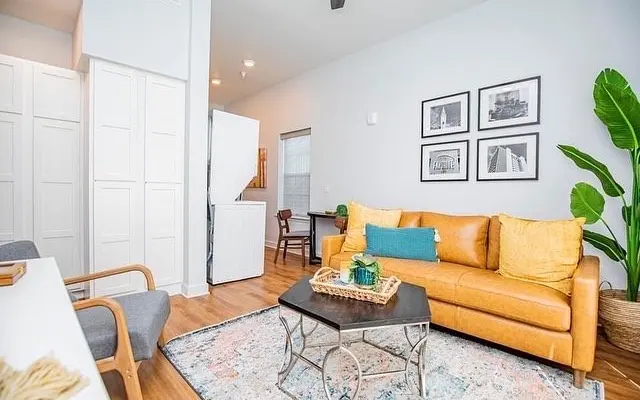 A modern living room featuring a leather sofa with decorative pillows, a coffee table with a tray, and framed artwork on the wall. There are potted plants and light wooden flooring in the space.