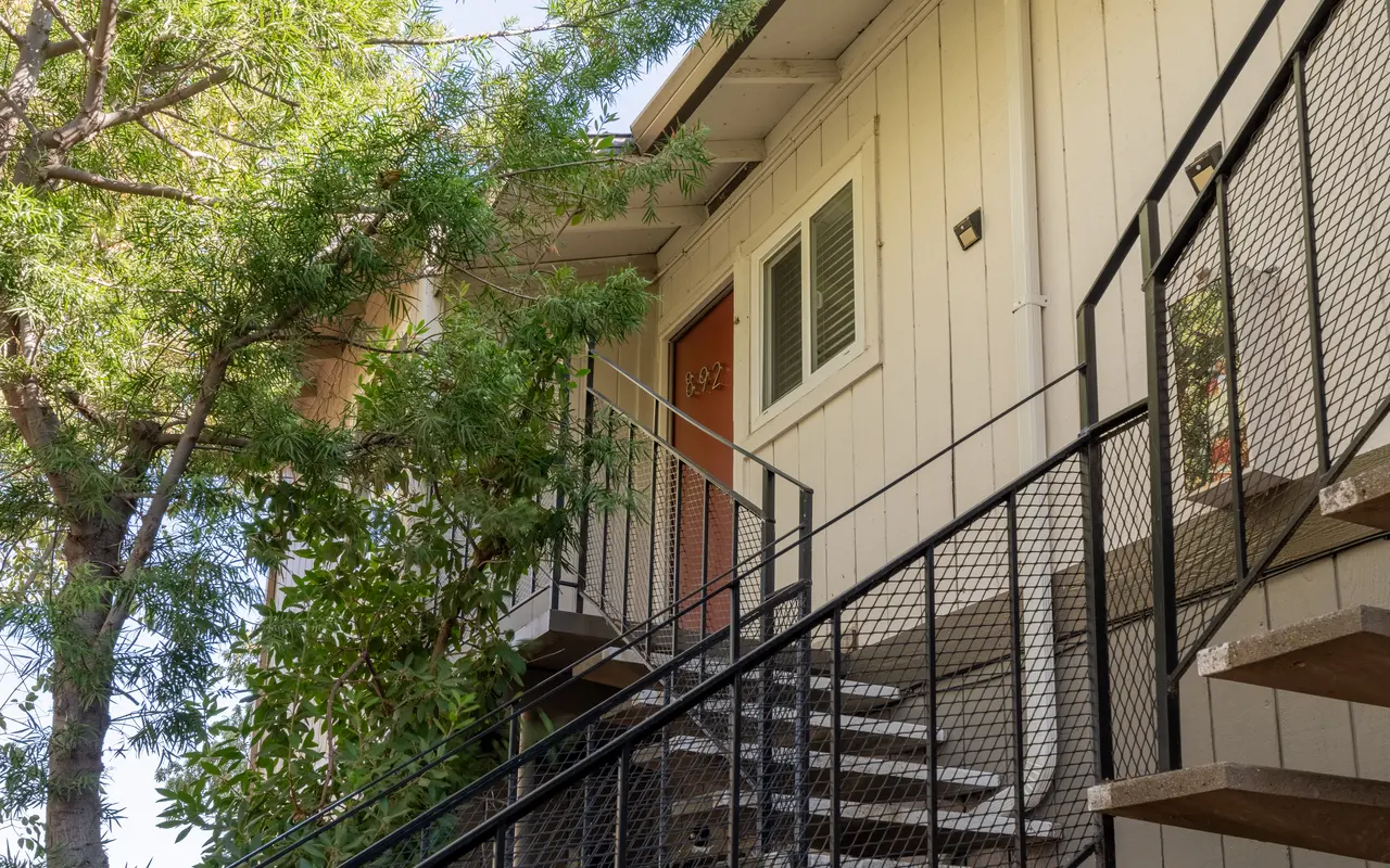 Exterior view of an apartment building with stairs leading to the entrance, surrounded by trees.