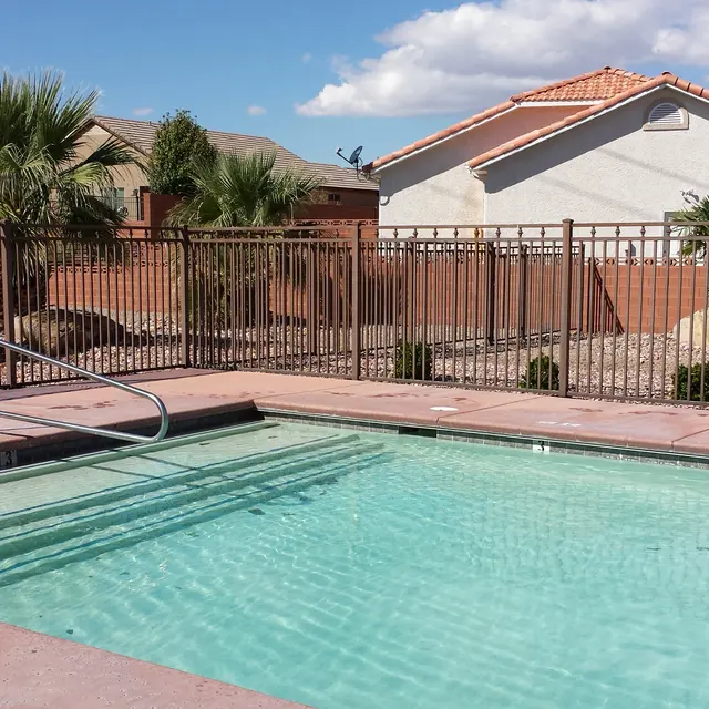 A swimming pool with a clear blue water, surrounded by a metal fence and framed by palm trees. In the background, there are residential houses and a patch of grass.