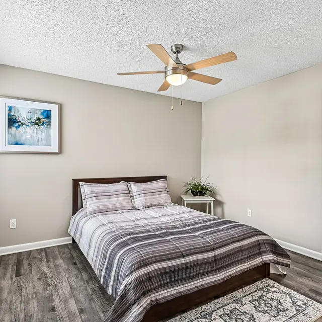 Modern Bedroom Interior A modern bedroom featuring a bed with a striped gray blanket and two white pillows. There's a ceiling fan above the bed, a small side table next to it, and a piece of artwork on the wall. Large windows with blinds allow natural light to brighten the room.