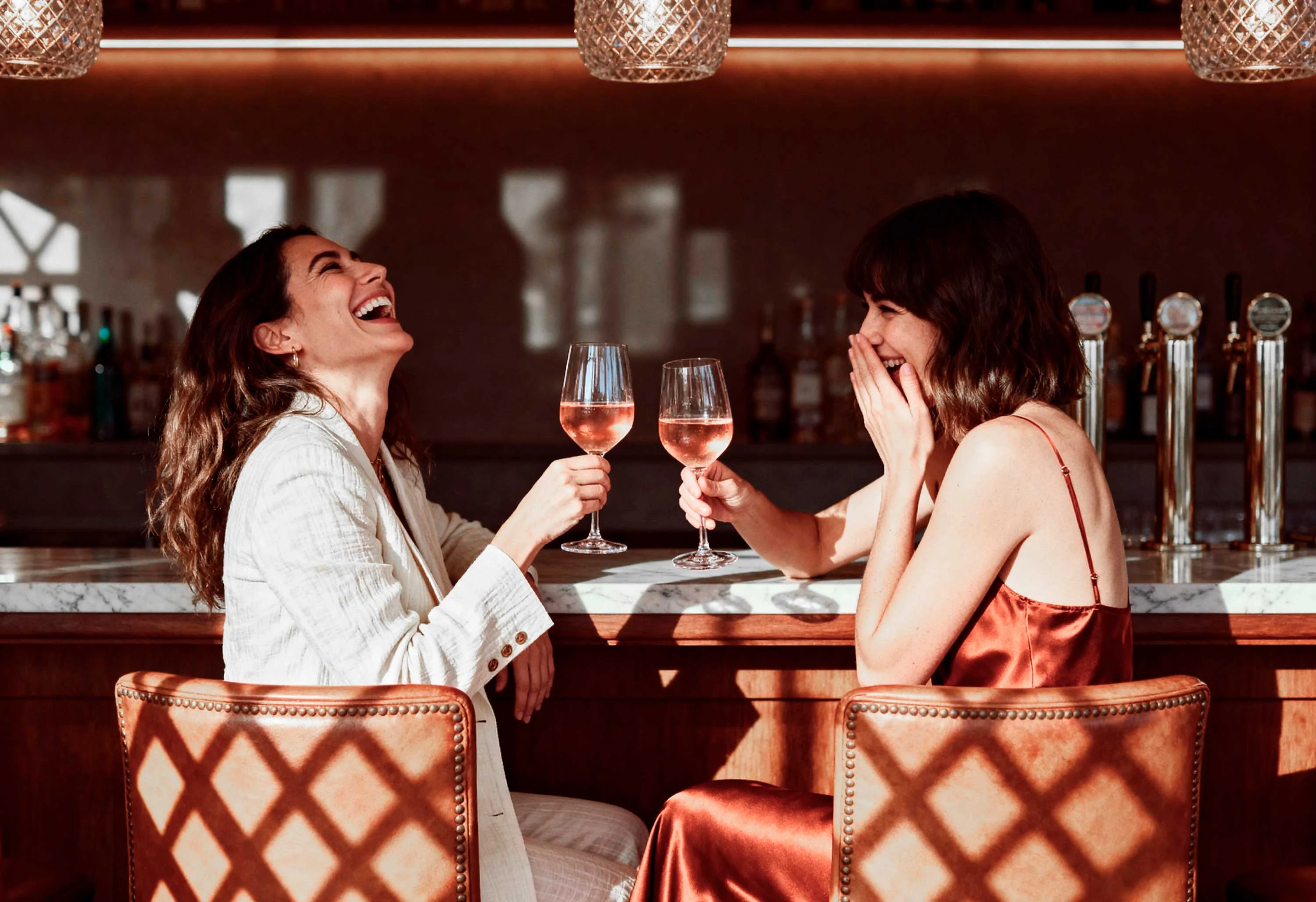 Joyful Toast at the Bar Two women laughing and toasting with wine glasses at a bar, surrounded by soft lighting and a stylish interior.