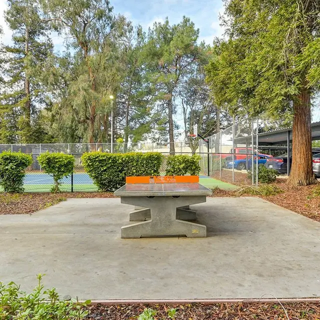 A concrete table with a bright orange center surrounded by greenery, with tennis courts in the background.
