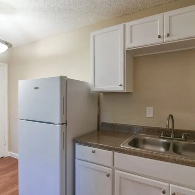 A tidy kitchen featuring white cabinets, a large silver refrigerator, and a double sink with chrome fixtures. The walls are a light beige color, and there is a ceiling light fixture providing illumination.