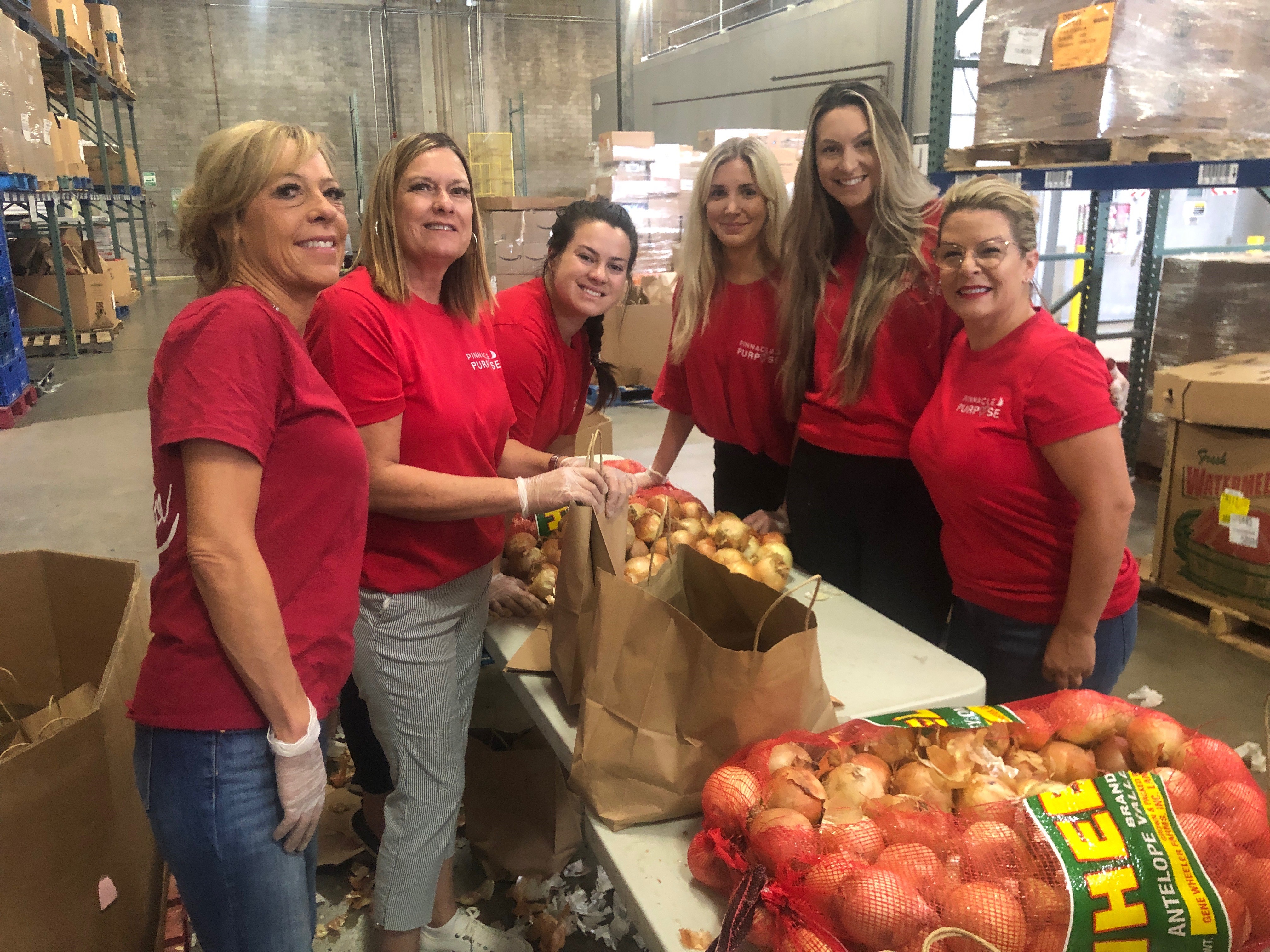 A group of six women in red shirts smiling and working together to pack onions in a warehouse.