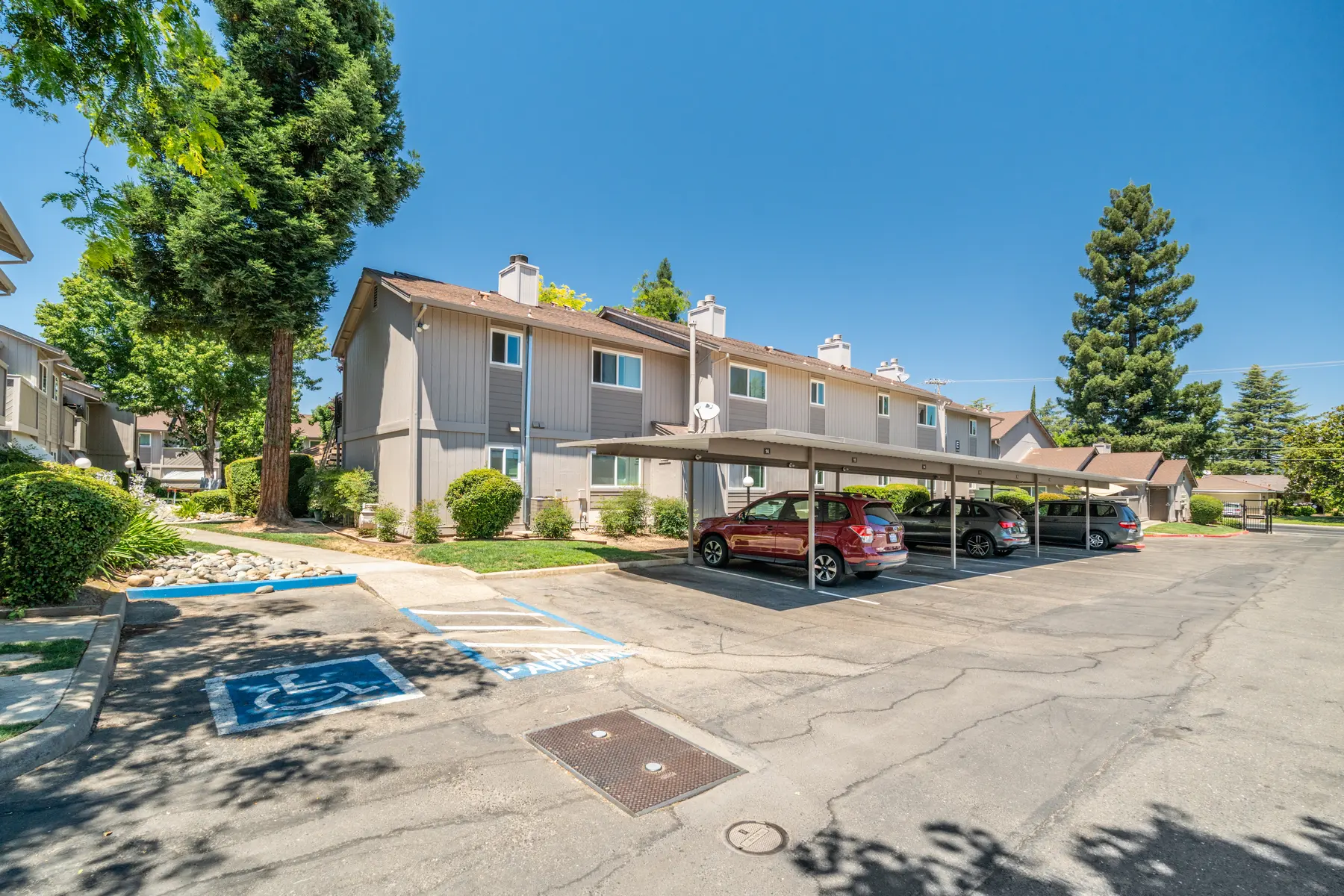 A view of an apartment complex featuring two-story buildings with parking spaces underneath a carport. The setting includes trees and clear blue skies.