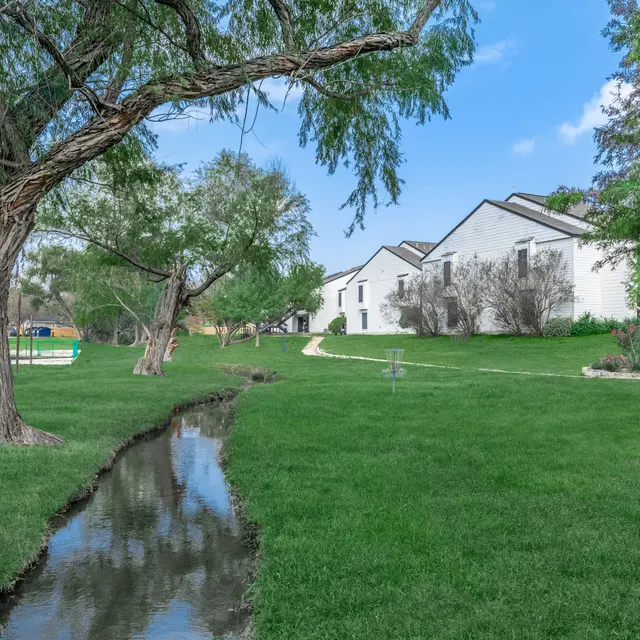 Grass area courtyard with a small stream running through it along with trees and a walkway. 