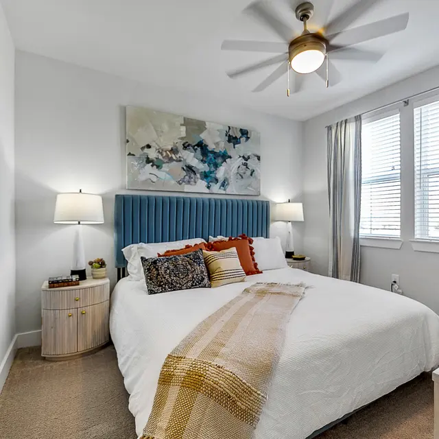 Cozy bedroom featuring a bed with colorful pillows, a patterned blanket, modern lamps, and large windows with curtains.