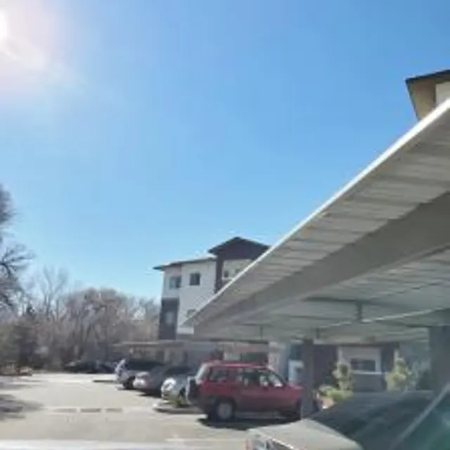 A parking lot view showing cars parked under a covered area with a building in the background against a clear blue sky.