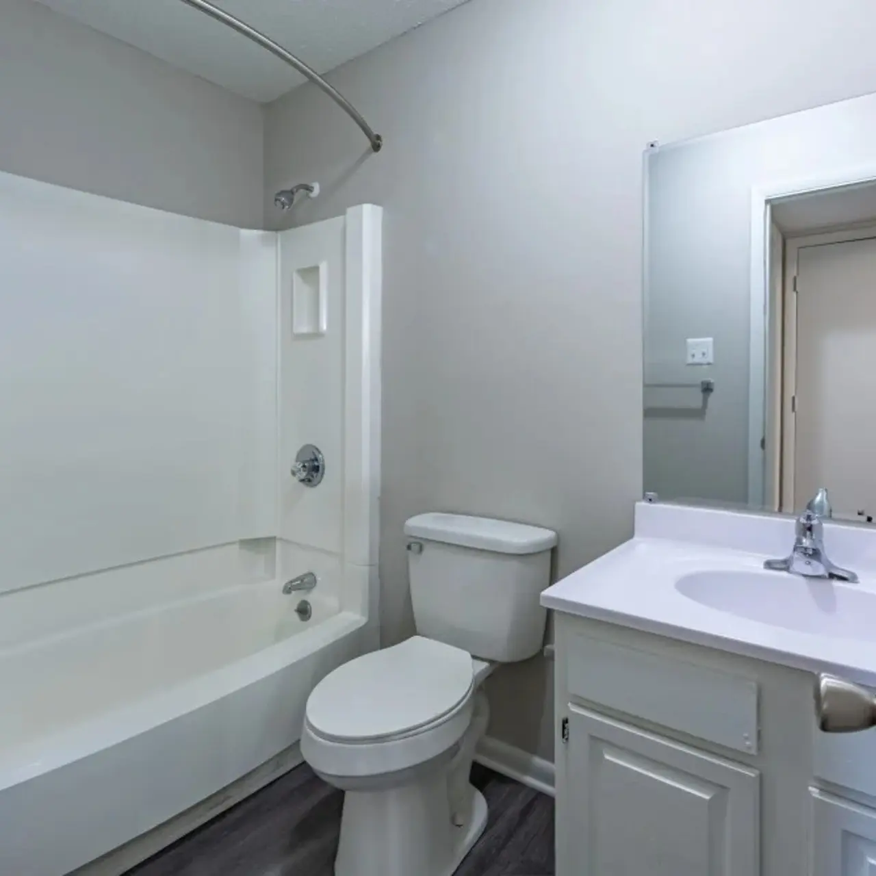 A clean and modern bathroom featuring a white bathtub, toilet, and sink with a mirror above the sink. The walls are painted in a light color, and the floor is a dark, wood-like material. There is a shower curtain rod above the bathtub, and a door leading to another room can be seen in the background.