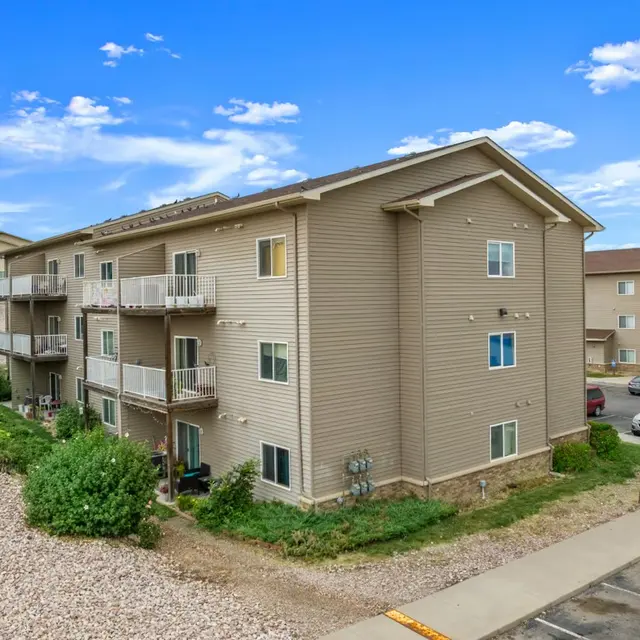 Eagle Ridge Three-story beige apartment building with balconies, surrounded by a gravel path and parking area, under a blue sky with clouds.
