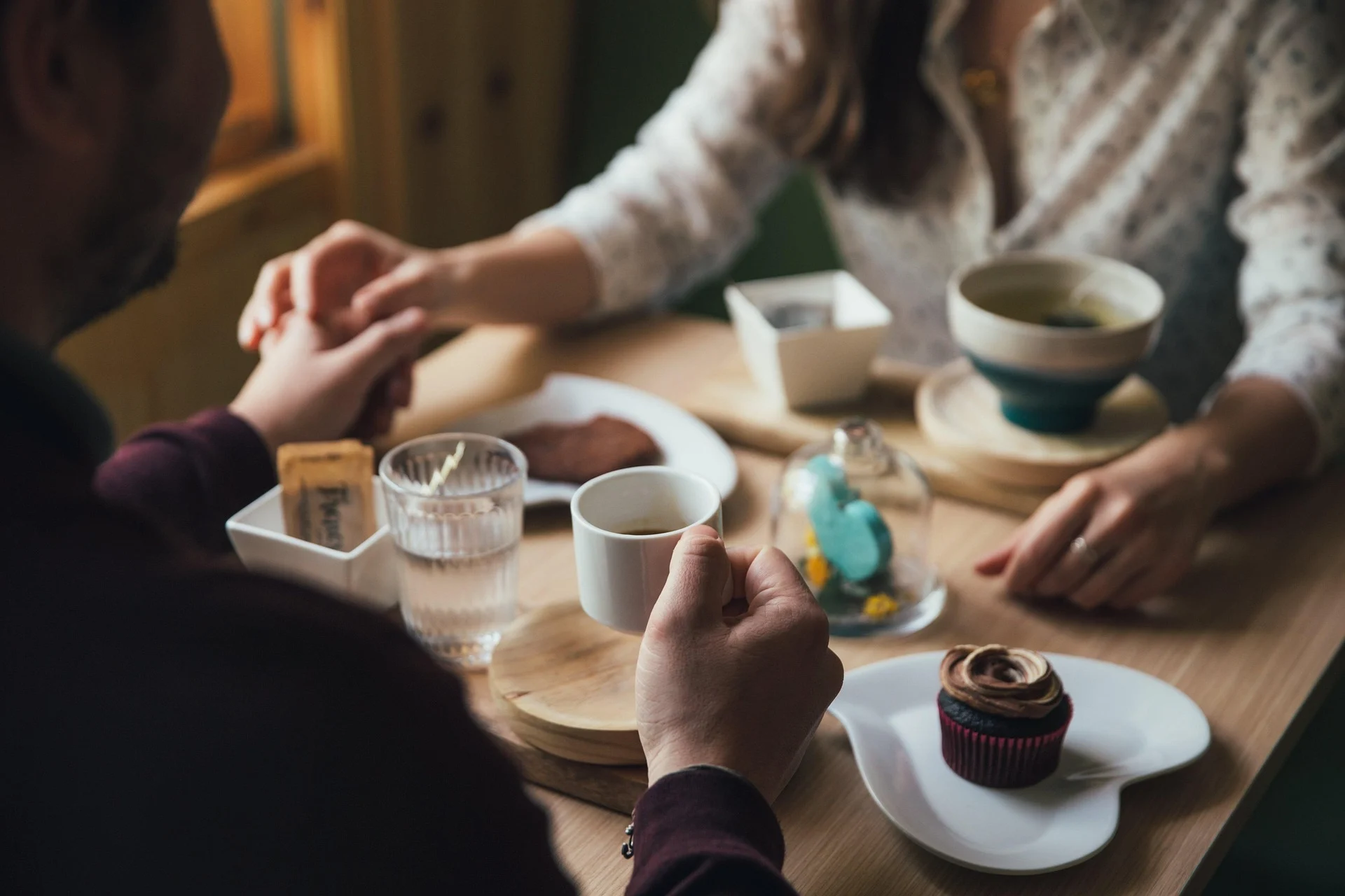 Cozy Morning Date Scene A cozy morning scene featuring two people holding hands across a table filled with drinks and desserts, including a cupcake and a bowl of soup, in a warmly lit environment.