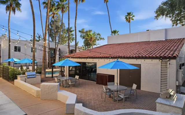 A cozy outdoor lounge area featuring a tiled roof building with several blue umbrellas over tables. Lush green palm trees surround the space, and string lights are visible in the background.