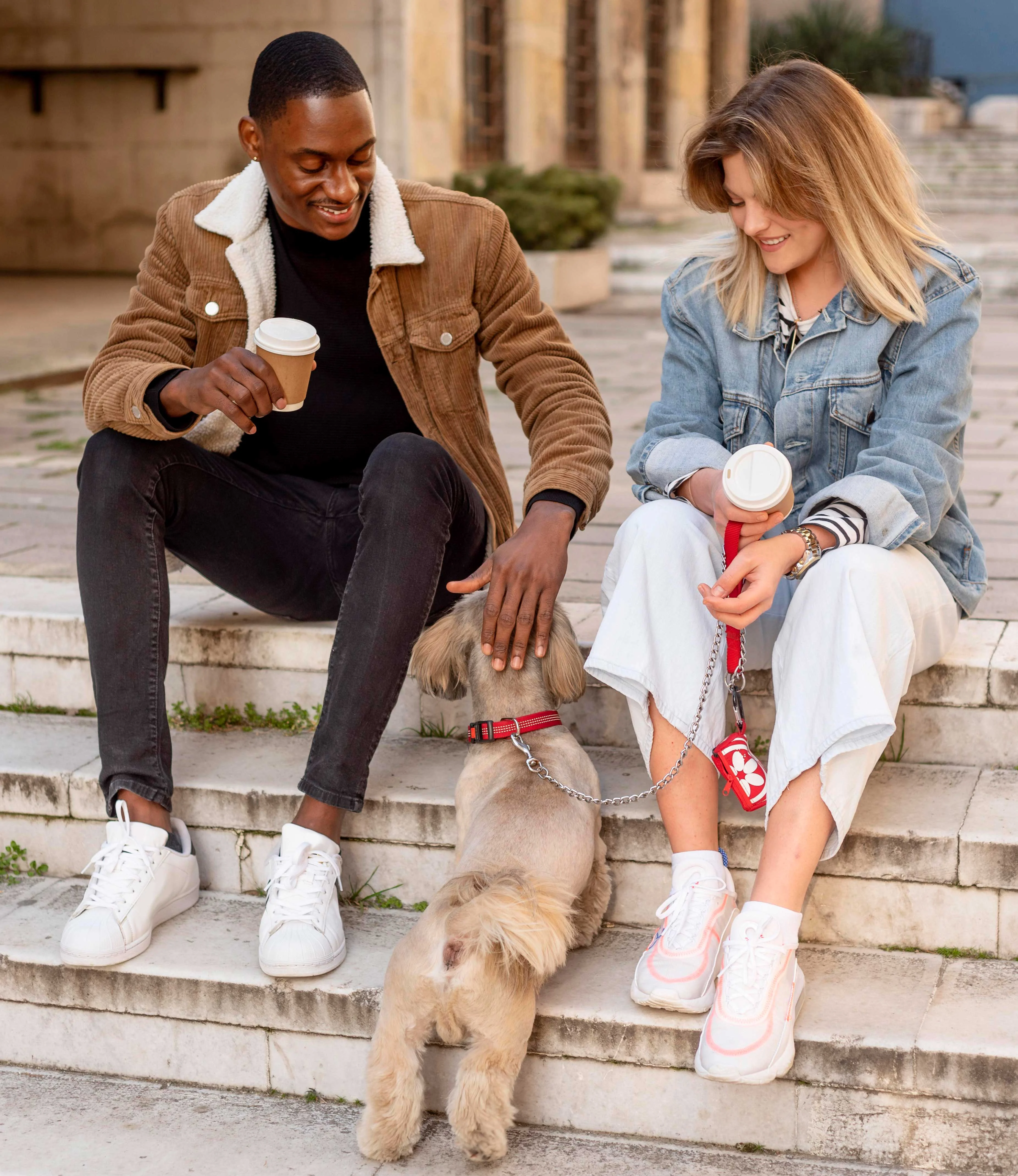 A couple sitting on steps with a small dog between them, both holding coffee cups.