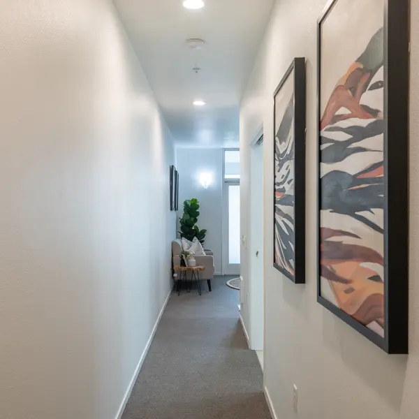 A narrow hallway with white walls, framed artwork on one side, and a small chair beside a potted plant at the end of the hallway.