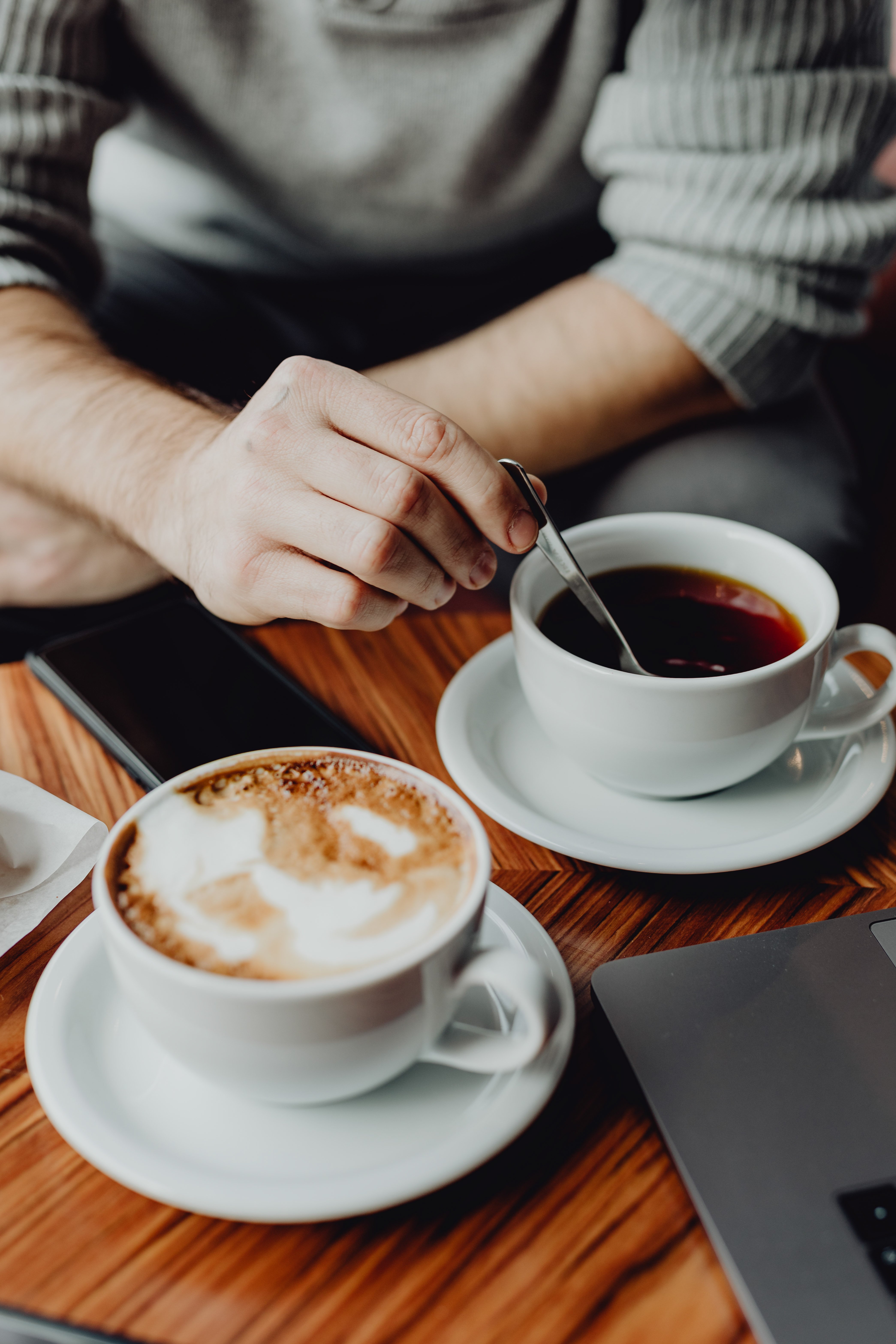 A pair of coffee cups on a wooden table with one person's hand stirring a cup. One cup contains cappuccino with foam, and the other is black coffee. There is a laptop nearby.