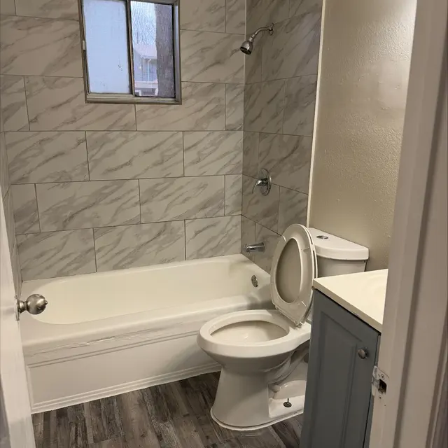 A modern bathroom featuring a shower bath with marble-style tiled walls, a toilet, and a sink cabinet with a gray finish. A small window allows natural light into the space.