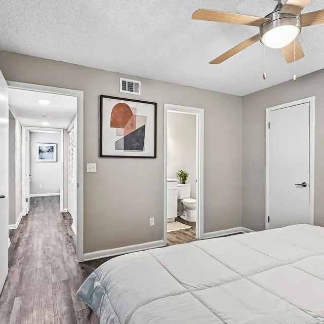 Modern Bedroom Interior A modern bedroom featuring a ceiling fan, a light-colored bedspread, and framed artwork on the wall. Two doors lead to different areas, one of which shows a bathroom. The room has light-colored walls and wooden flooring.
