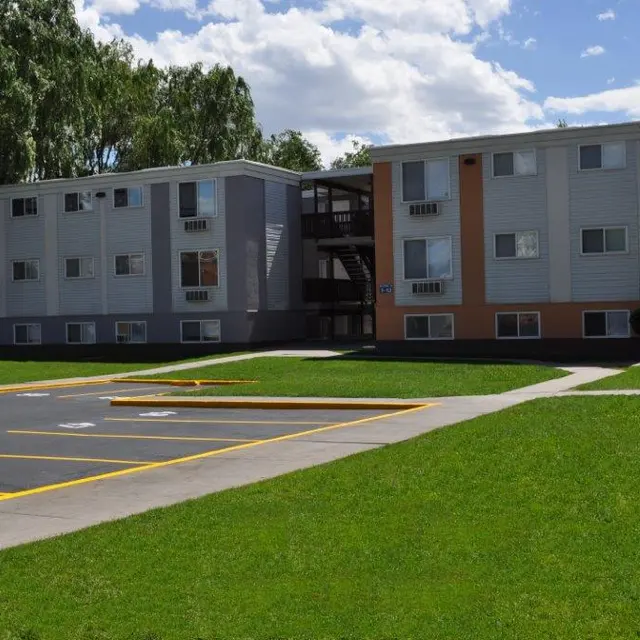 A view of a multi-story apartment building with a green lawn and parking spots.