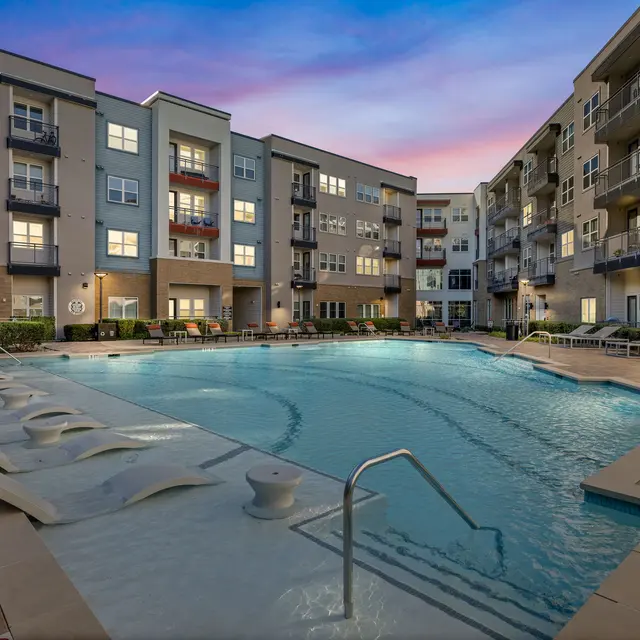 A modern apartment complex featuring a large swimming pool surrounded by lounge chairs and balconies overlooking the area, bathed in a sunset glow.