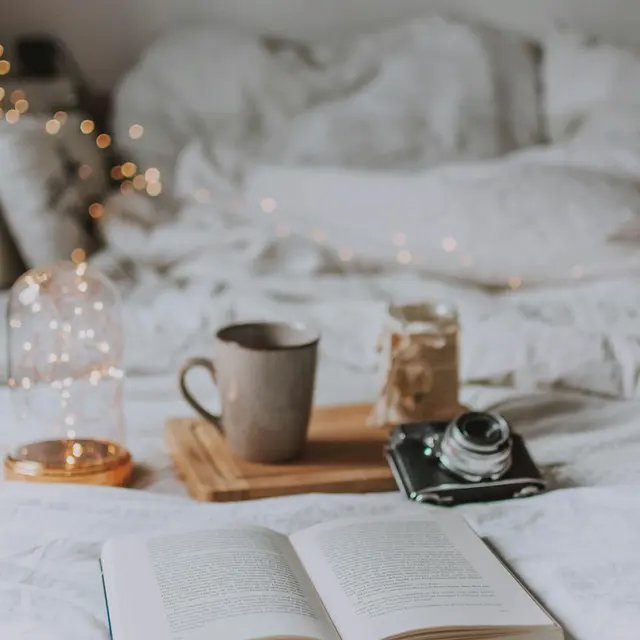 A cozy bedroom setup featuring an open book on a bed, a cup of coffee, a jar, and a camera on a wooden tray. Soft bedding and fairy lights create a warm atmosphere.