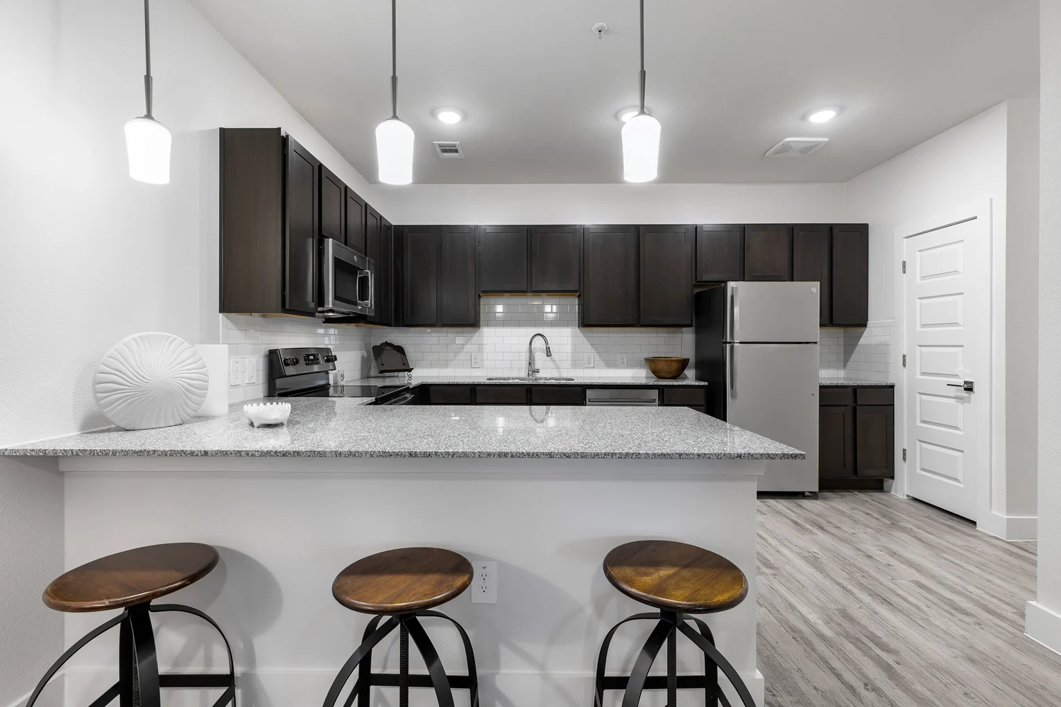 A modern kitchen with dark cabinetry and stainless steel appliances. The kitchen island features three wooden stools and is adorned with a decorative item and a bowl.