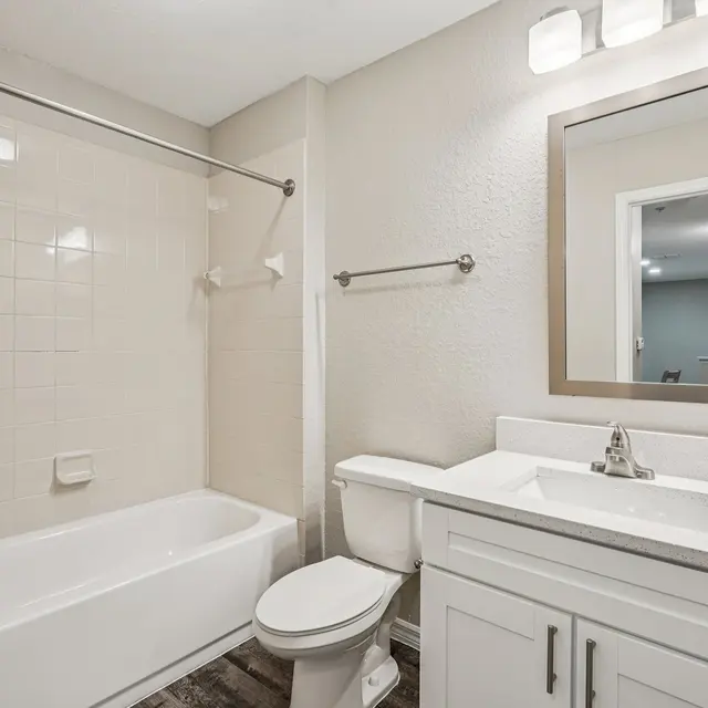 A clean and modern bathroom featuring a bathtub, a toilet, and a sleek vanity with a mirror. The walls are light-colored, and the floor is designed with wood-like tiles.