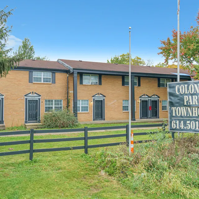 Front view of Colonial Park Townhomes with a sign and well-maintained landscaping, featuring green grass and colorful trees in the background.