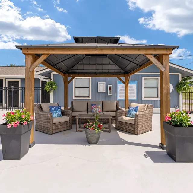 An outdoor lounge area with a wooden gazebo featuring wicker furniture and potted flowers, set against a blue sky and a building in the background.