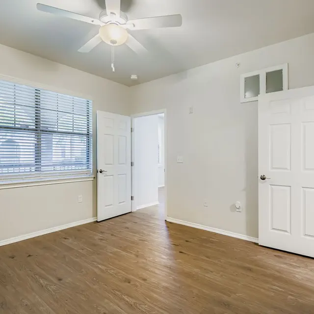 An empty room with a wooden floor, a ceiling fan, and two open doors. There are two windows with white blinds on one wall.