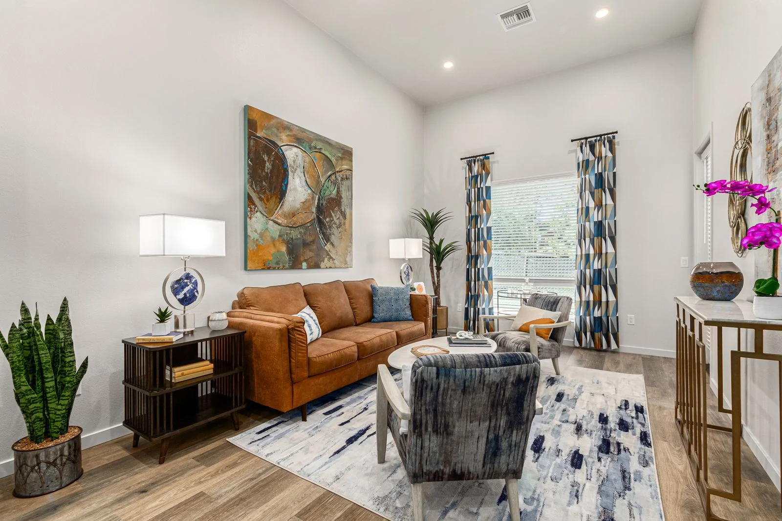 Modern Living Room Decor A stylish modern living room featuring a brown sofa, decorative pillows, a patterned rug, and artistic wall decor. There are plants and decorative items on display, along with curtains and natural light flowing through the window.