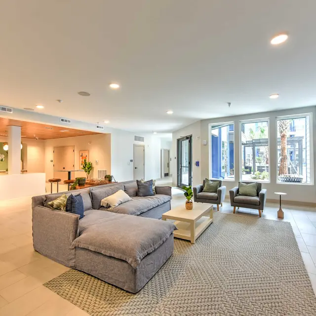 Spacious Modern Living Area A spacious modern living area with a large sectional couch, a coffee table, and two armchairs. Windows allow natural light to enter, showcasing a cozy and inviting atmosphere. The decor features neutral tones and a textured rug, complemented by a decorative wall.