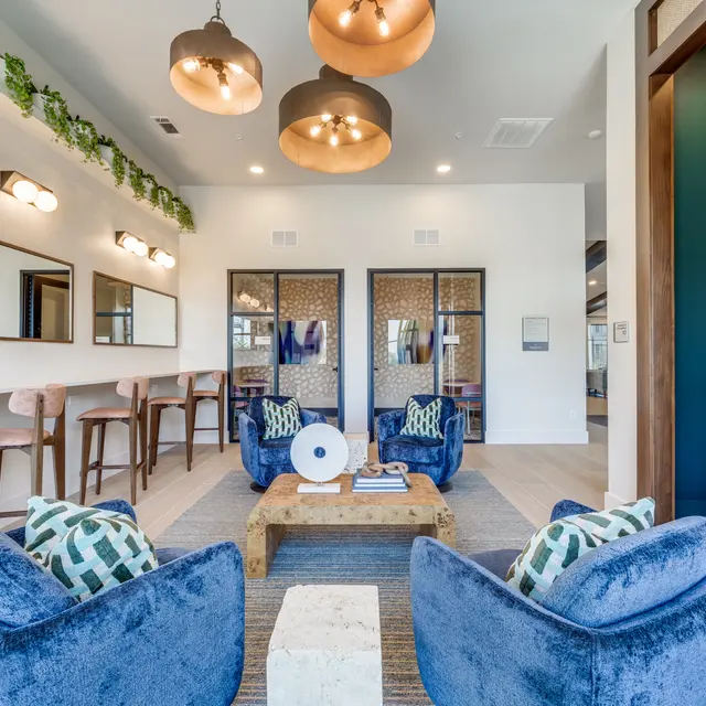 Modern living area featuring two plush blue chairs, a wooden coffee table, and a bar-style seating arrangement along the back wall; illuminated by stylish hanging lights and decorated with greenery.