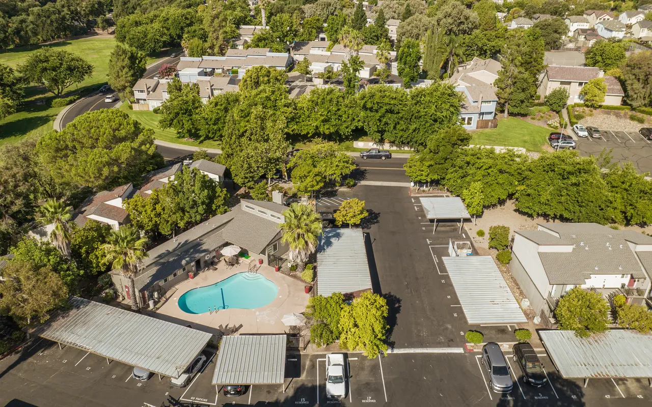 Aerial view of an apartment complex featuring a swimming pool surrounded by trees, with residential buildings visible in the background.