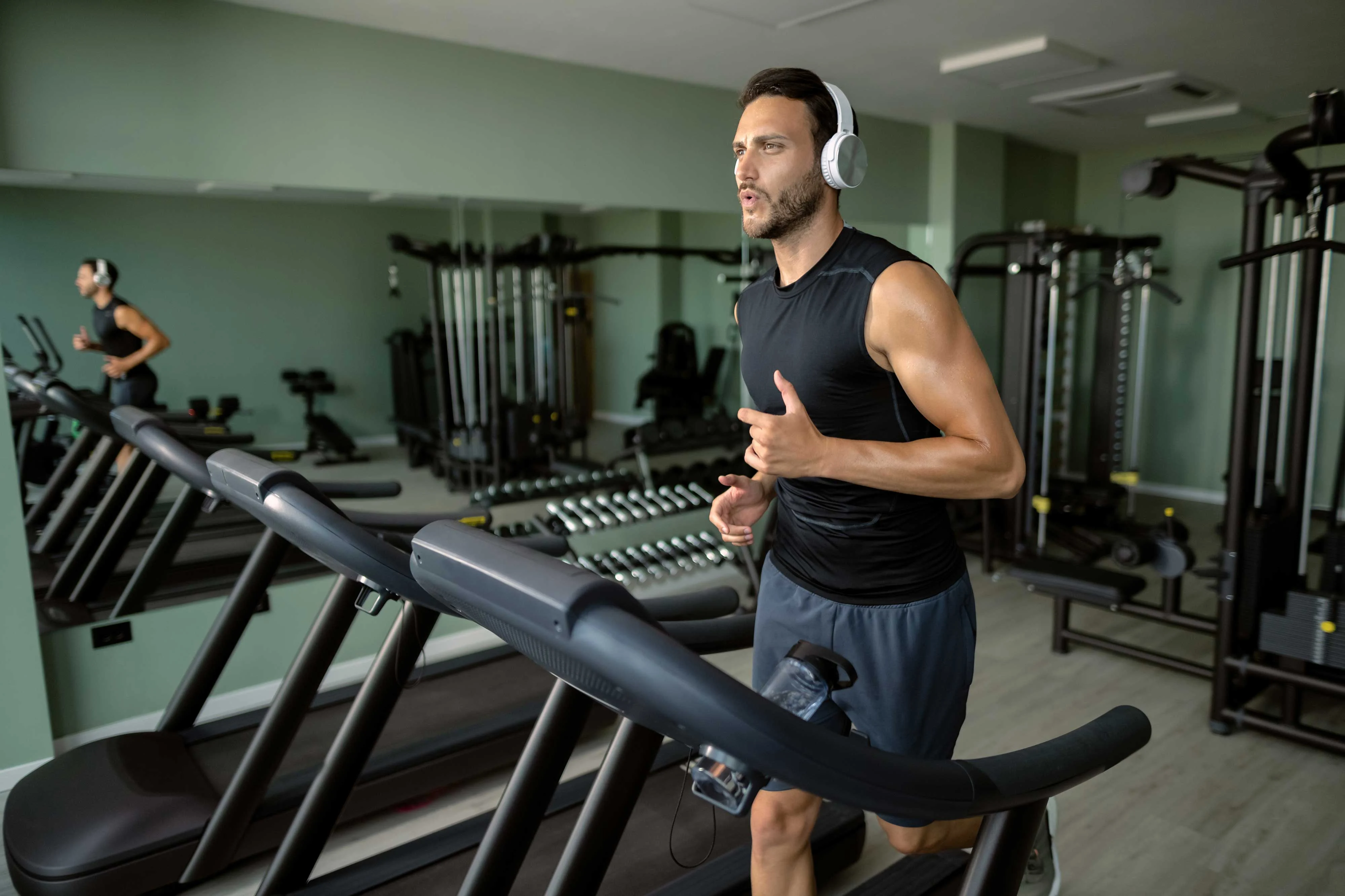 A man jogging on a treadmill in a modern gym, wearing headphones and athletic gear, with gym equipment in the background.