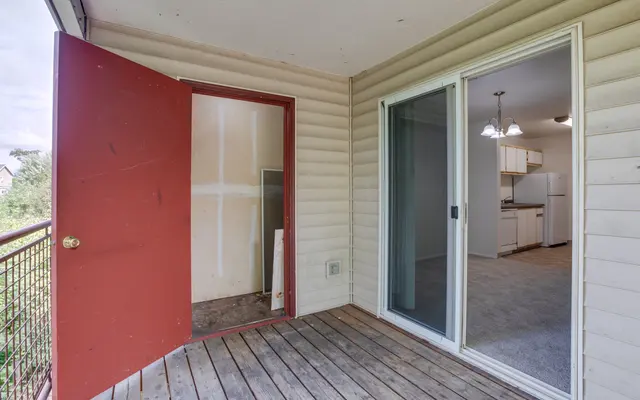 A balcony with a red door leading to a storage space and a sliding glass door that opens into an interior room. The balcony has wooden flooring and a light-colored exterior wall.