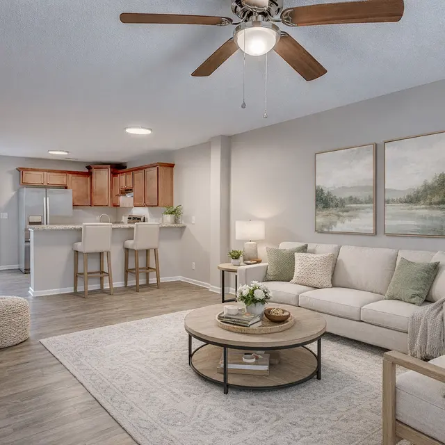 Contemporary Living Room with Kitchen Access A cozy living room featuring a light sofa, coffee table, and a flat-screen TV. The room has a ceiling fan and is connected to a kitchen area with wooden cabinets. Large windows allow natural light to fill the space.