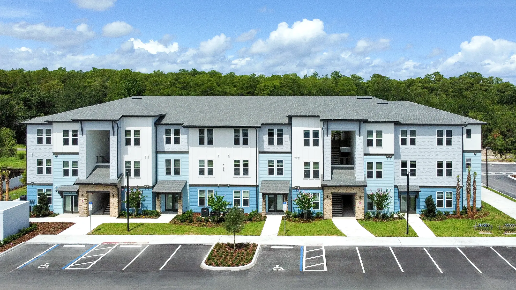 A modern three-story apartment complex with a light blue and gray exterior, surrounded by green landscaping and a parking lot.