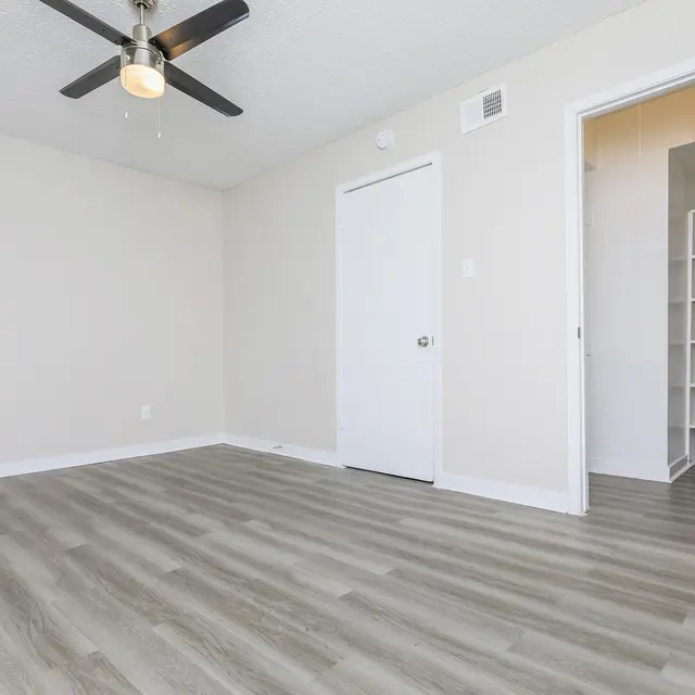 A spacious empty room with light wood flooring, a ceiling fan, and a white door leading to a small closet. The walls are painted light beige.