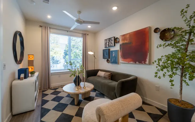 A modern living room with a green sofa, patterned rug, and natural light coming from the window. There are decorative elements like artwork on the walls and a faux plant in the corner.