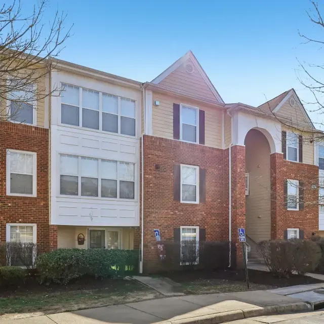 Exterior view of a brick apartment building with multiple windows and an entrance archway, surrounded by grass and shrubs.