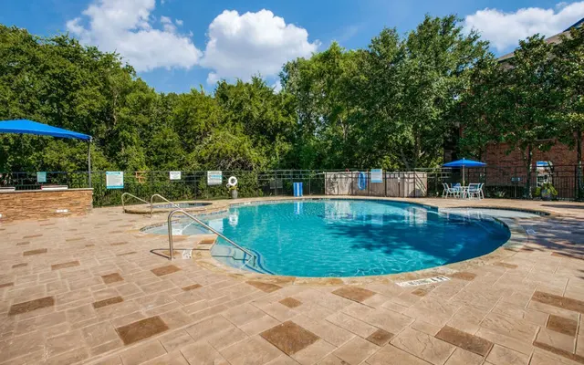 A clear swimming pool surrounded by beige tiled deck, with blue umbrellas and greenery in the background.