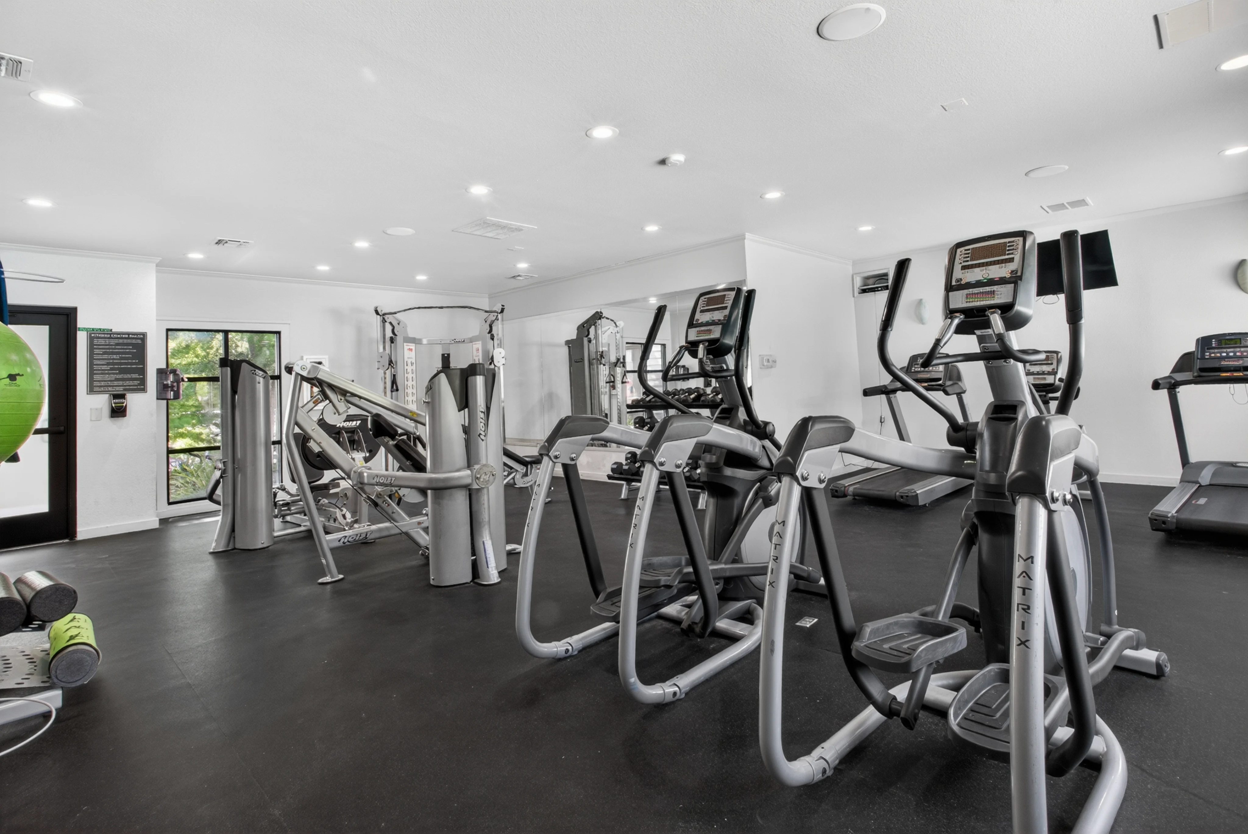 Interior view of a modern gym featuring various fitness equipment including ellipticals and treadmills, with a large window allowing natural light in.