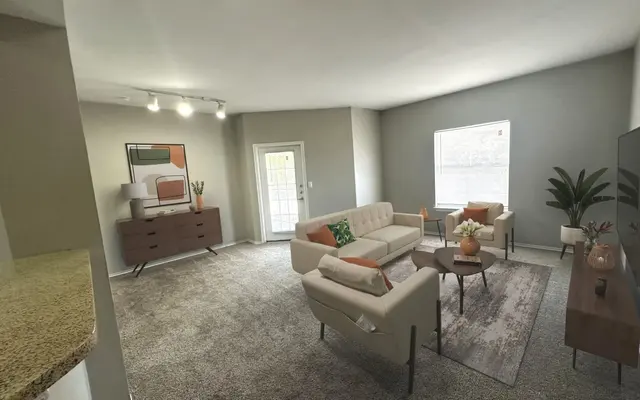 A spacious modern living room with a large window, light gray walls, and plush carpet. The room features a white sectional sofa, two cream armchairs, and a brown wooden side table with decorative items. A flat-screen television is mounted on a console across from the seating area, and a potted plant adds a touch of greenery. The decor is minimalist and stylish, making the space feel inviting and bright.