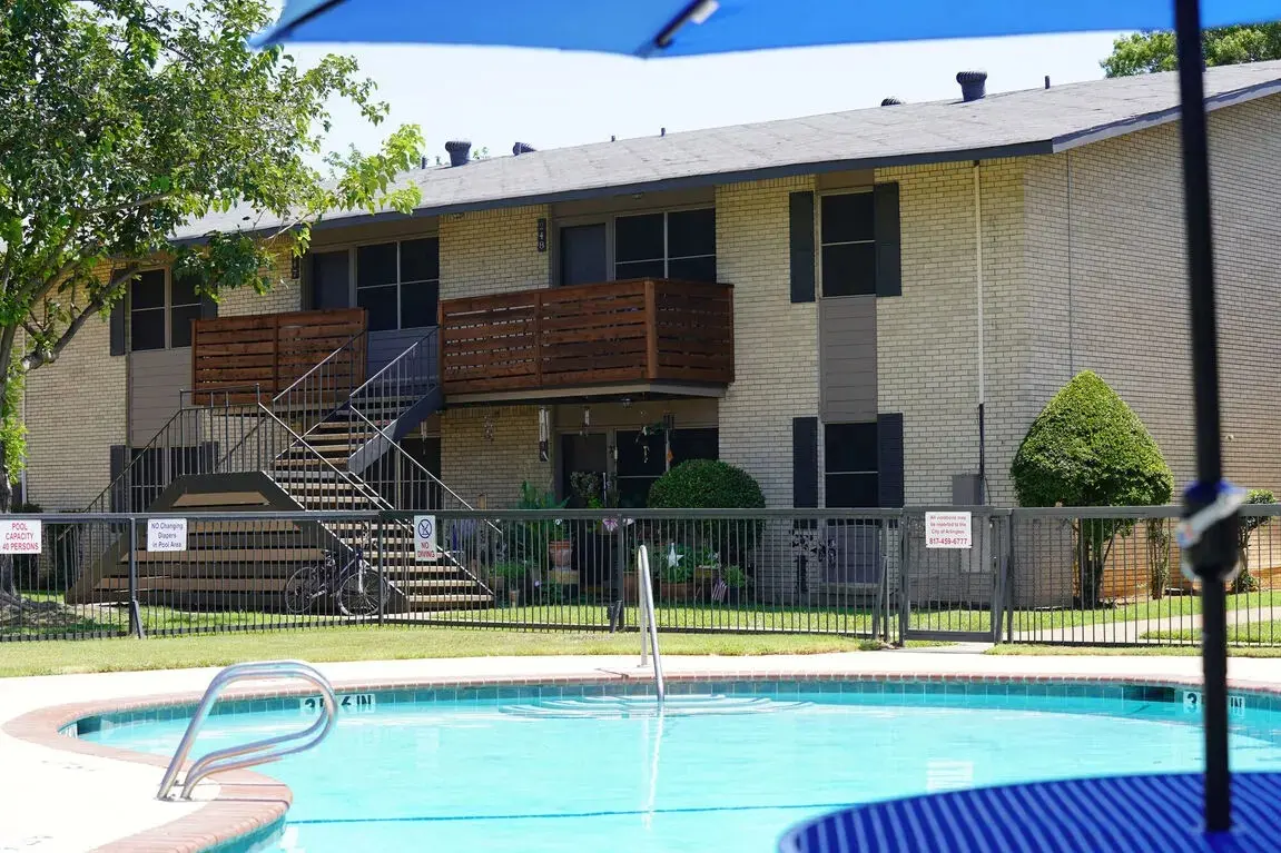 View of a swimming pool in front of an apartment building, featuring two balconies and landscaped greenery.