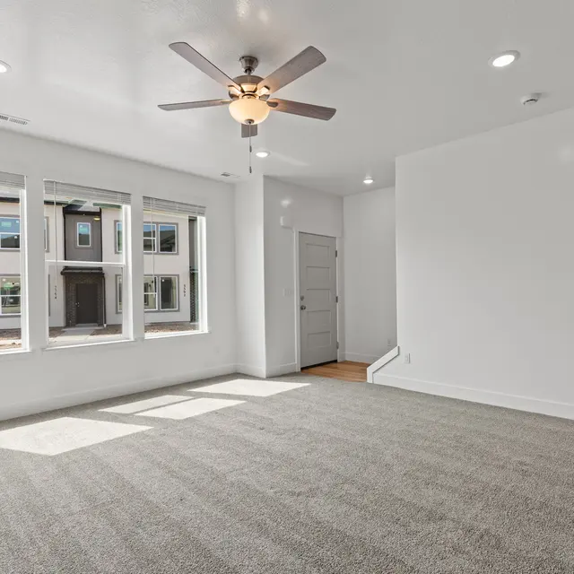 An empty living room with gray carpet, ceiling fan, and large windows facing outside. A door leads to another area, and there is a white wall and minimal furniture in the space.